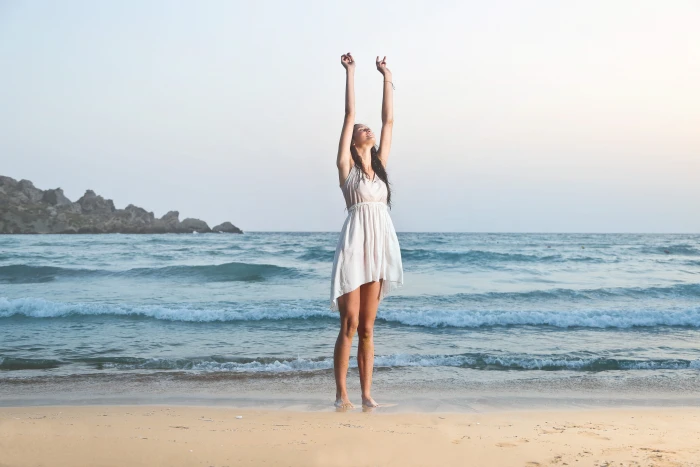 A young brunette woman wearing a white dress standing on the beach with her hands up 2k