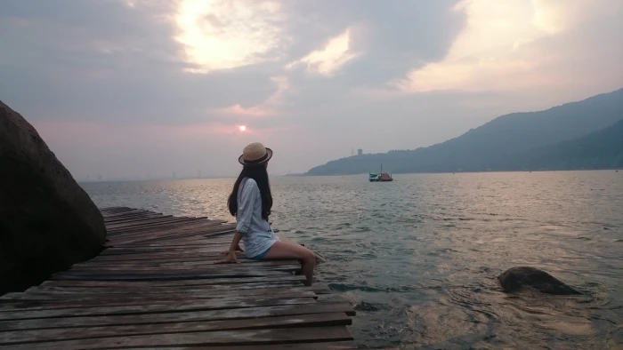 woman wearing white long sleeved top sitting on brown wooden dock facing body of water 2k 4k
