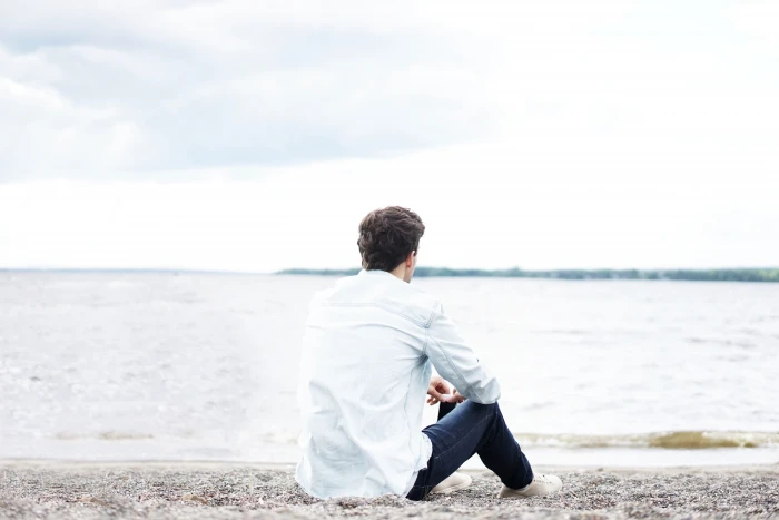 man in white dress shirt and black denim jeans sitting on sand watching seawave during daytime 2k 4k 5k