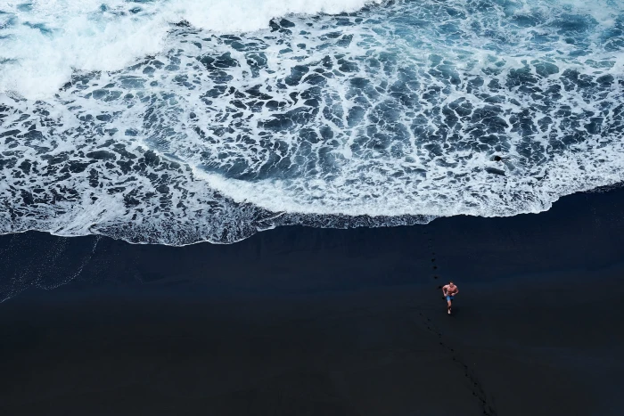 person walking on black sand near body of water beach men white 2k