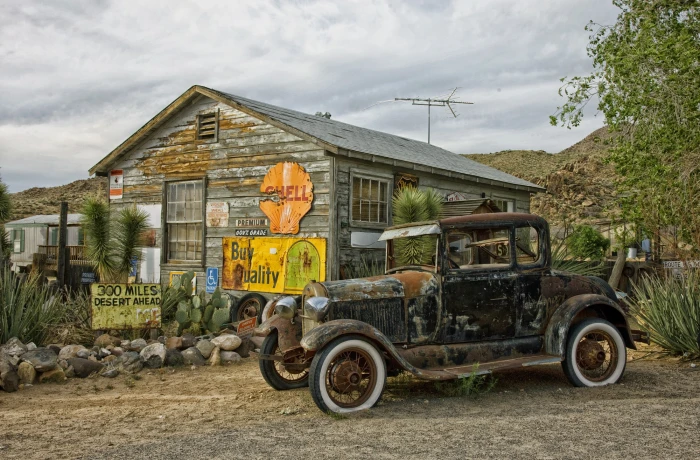 rusted vintage black and brown vehicle near white wooden house under cloudy blue sky at daytime 2k
