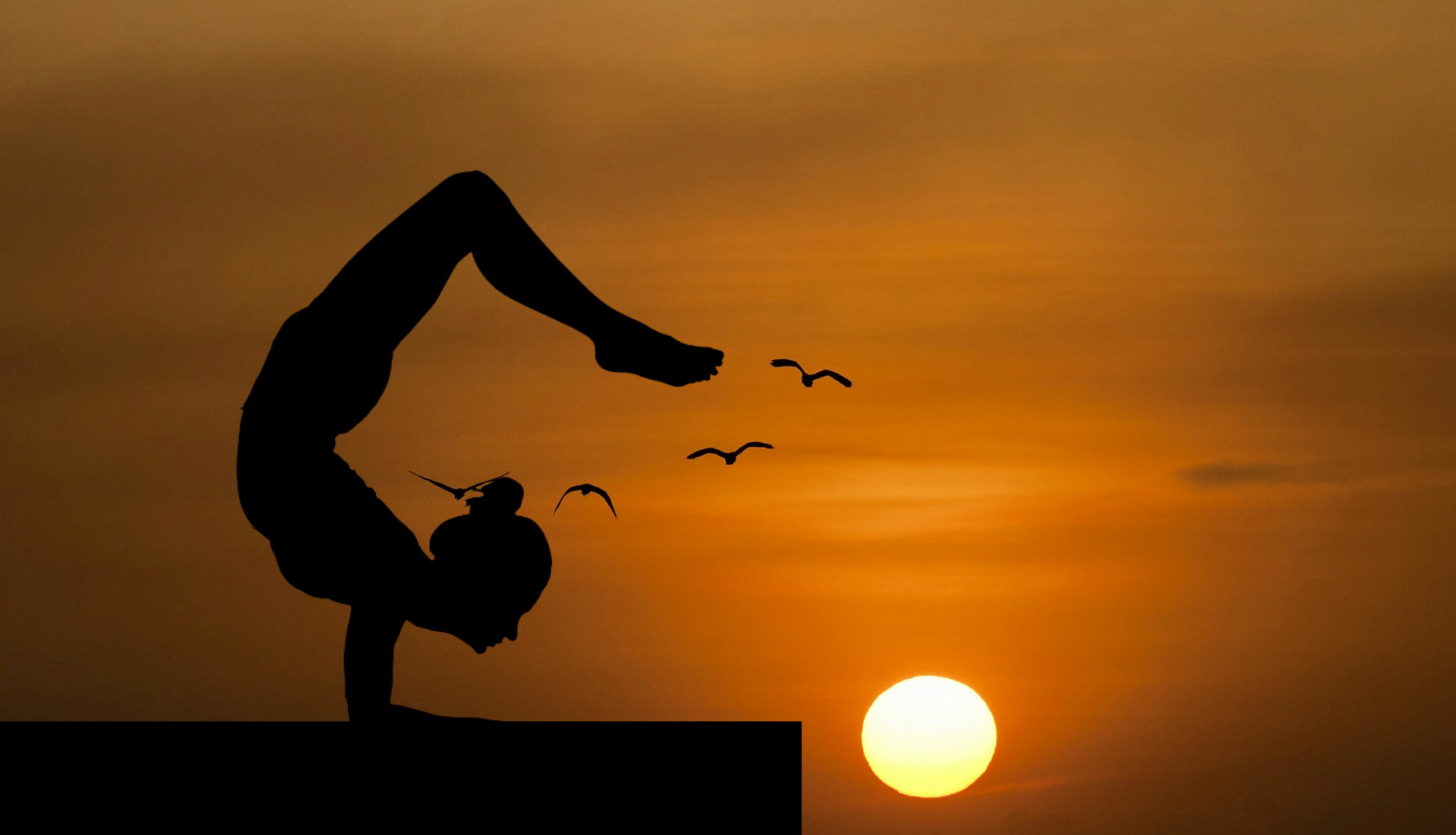 Woman doing an acrobatic yoga pose silhouetted against sunset 2k