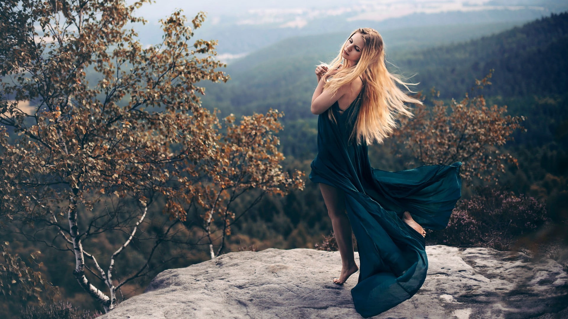 women's blue sleeveless slit dress woman wearing teal posing with both of her hands on heart while she close eyes grey cliff surrounded brown trees 2k