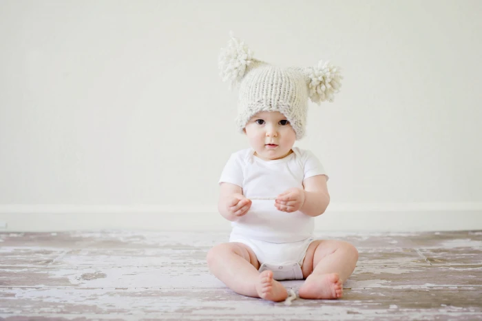baby wearing white onesie with beige bubble cap sitting on the floor 2k 4k