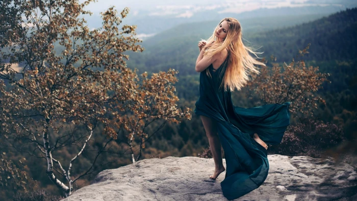 women's blue sleeveless slit dress woman wearing teal posing with both of her hands on heart while she close eyes grey cliff surrounded brown trees 2k