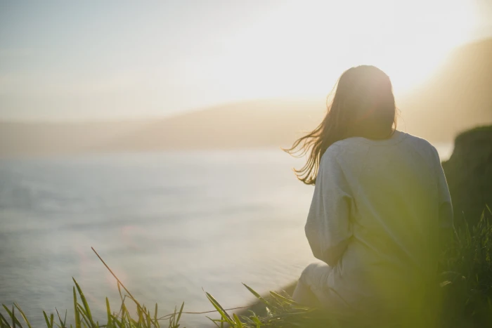 woman wearing gray long sleeved shirt facing the sea sitting on cliff during daytime 2k 4k