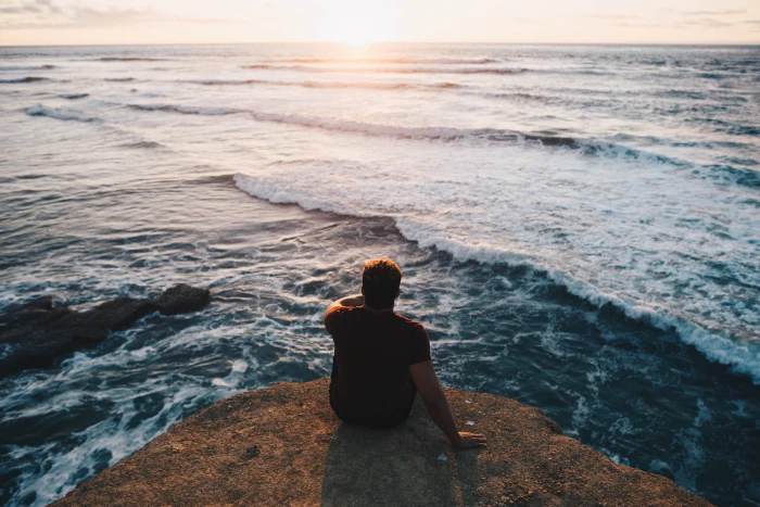 person sitting on cliff facing body of water man rock near sea 2k 4k 5k
