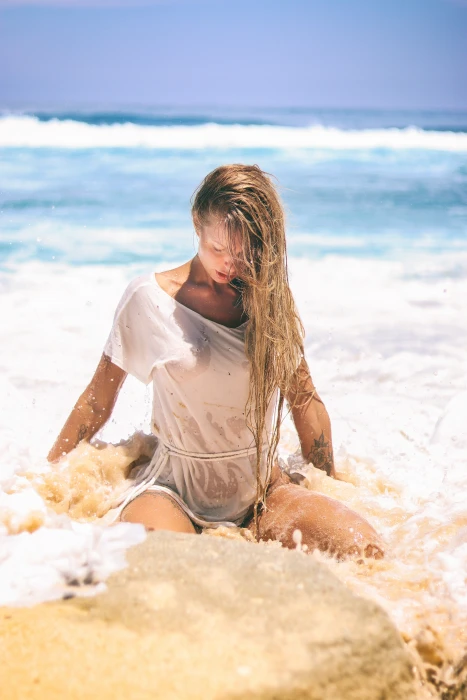 woman in white top sitting on sand near body of water selective focus seashore during daytime 2k 4k